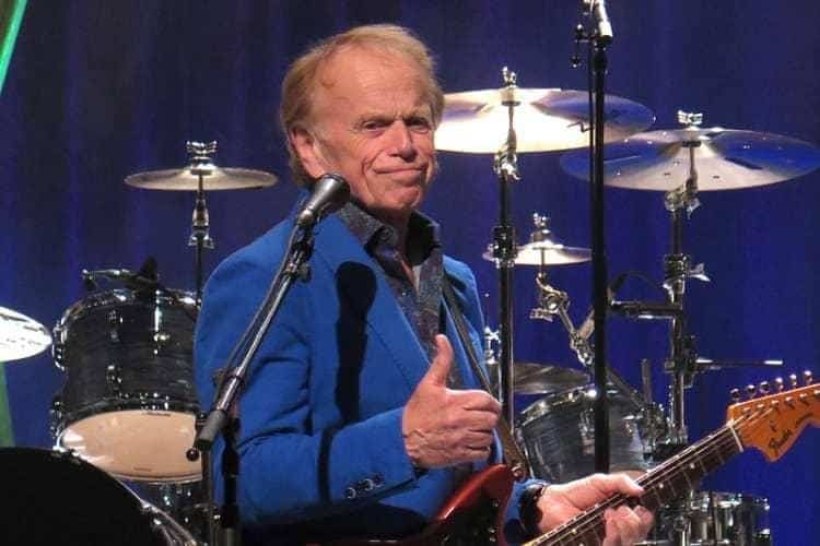 Musician in blue shirt playing guitar and singing at microphone, drum kit visible behind on stage.