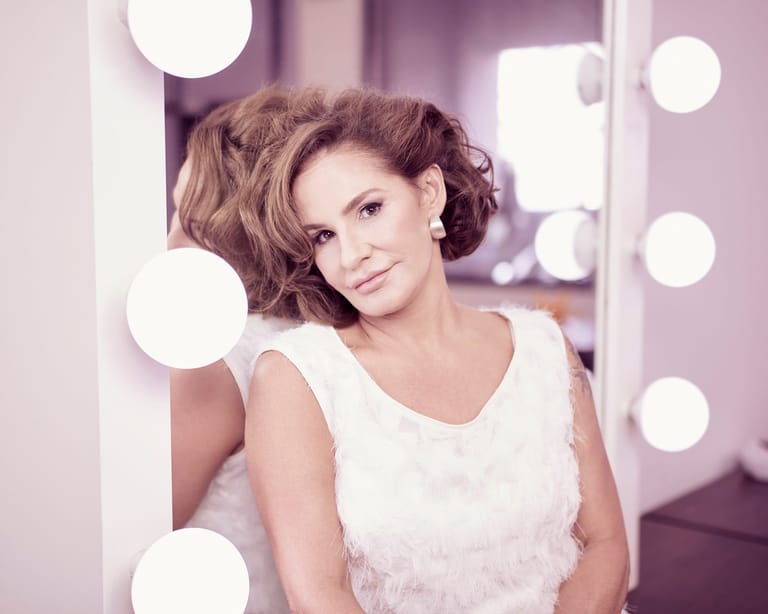 Woman in white sleeveless top seated before illuminated vanity mirror with soft purple-toned backdrop.