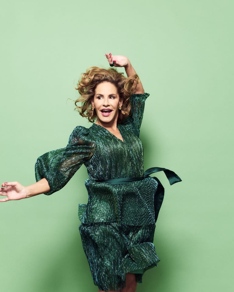 Woman in a shimmering green jacket poses playfully against a pale green background, hand raised to her head.