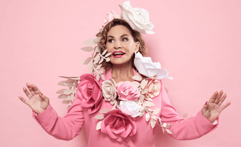 Woman in pink outfit adorned with oversized flowers against a pink background, arms raised expressively.