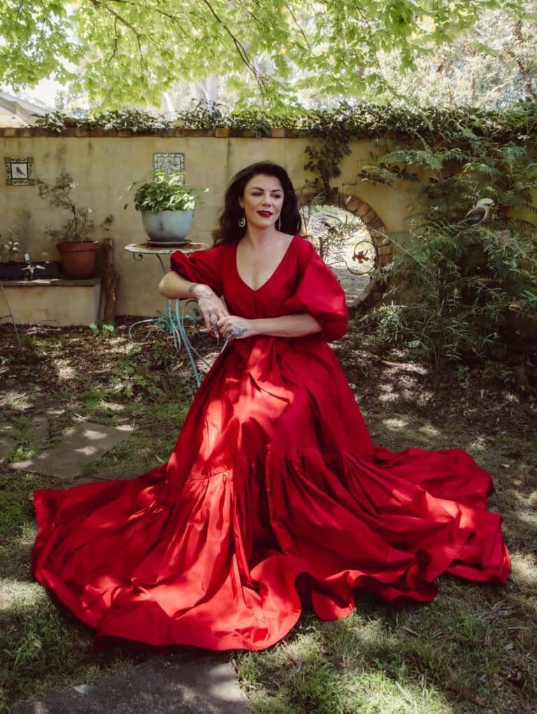 Woman in a flowing red gown seated in a sunlit courtyard garden surrounded by climbing vines and potted plants.