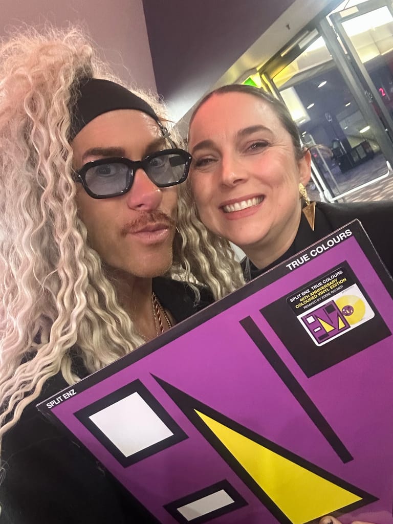 Two people with long blonde wigs pose together holding a bright purple and yellow True Colours album artwork.