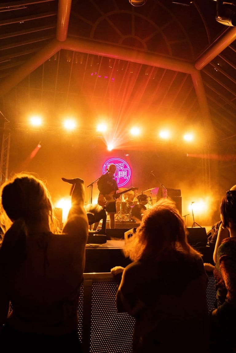 Band performing on stage bathed in warm orange lighting with neon pink logo, audience silhouettes in foreground.