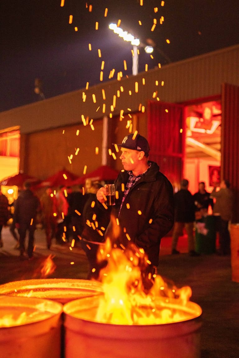 Performer holds microphone whilst standing before an open fire with sparks exploding upward in an outdoor nighttime venue.