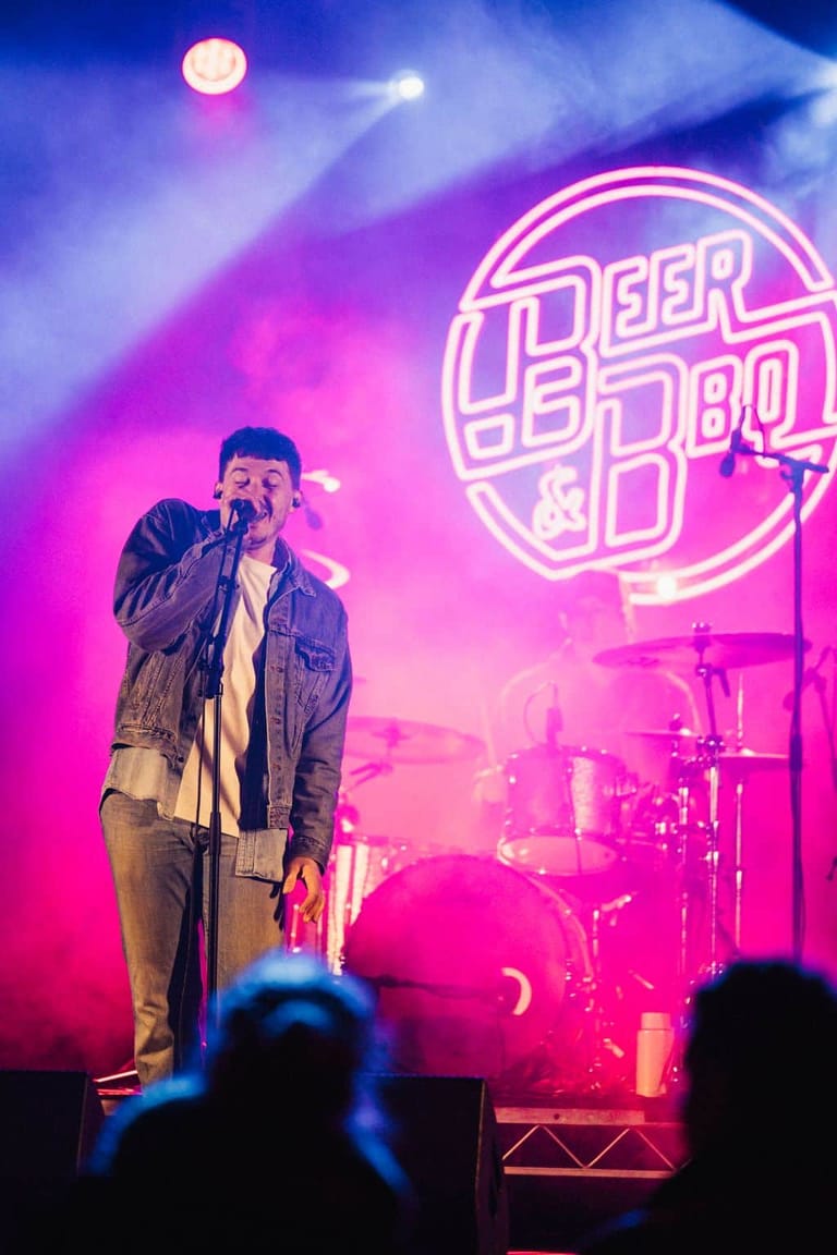 Male performer singing into microphone on stage beneath neon signage, bathed in pink and blue stage lighting with drummer visible behind.