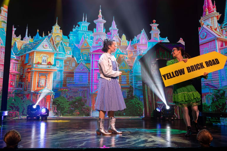 Singer in vintage dress performs centre stage before an illuminated fairy-tale castle projection during a theatrical tour production.