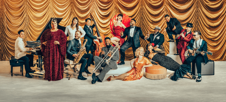 Large ensemble of musicians and performers in formal attire posed against ornate golden draped backdrop.