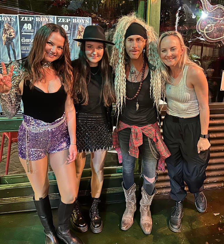 Four people pose together in a venue with neon signage, wearing casual and rock-inspired outfits.