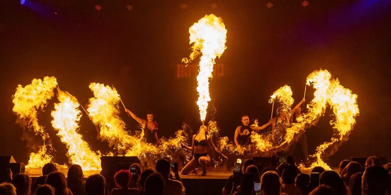 Multiple fire breathers exhale flames in synchronised arcs above a stage, with silhouetted audience members watching in the foreground.