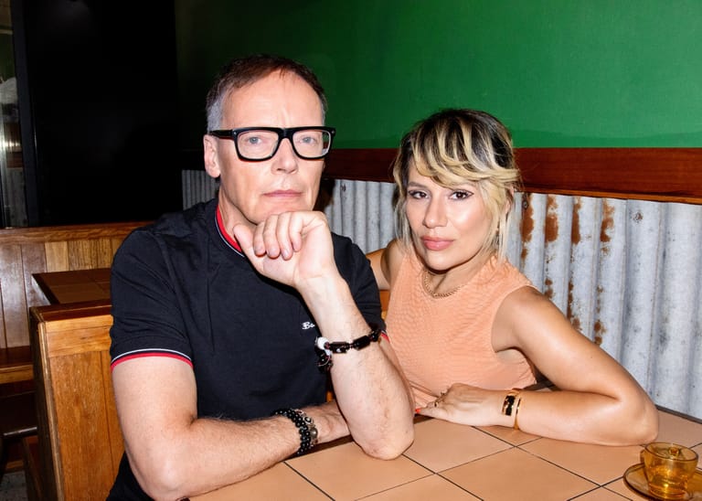 Two people seated at a table against a green wall, one wearing glasses and black polo shirt, the other in a peach-coloured top.