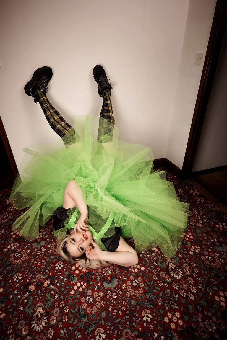 Performer in a vibrant lime-green tutu lies on a patterned floor with legs raised theatrically against a white wall.