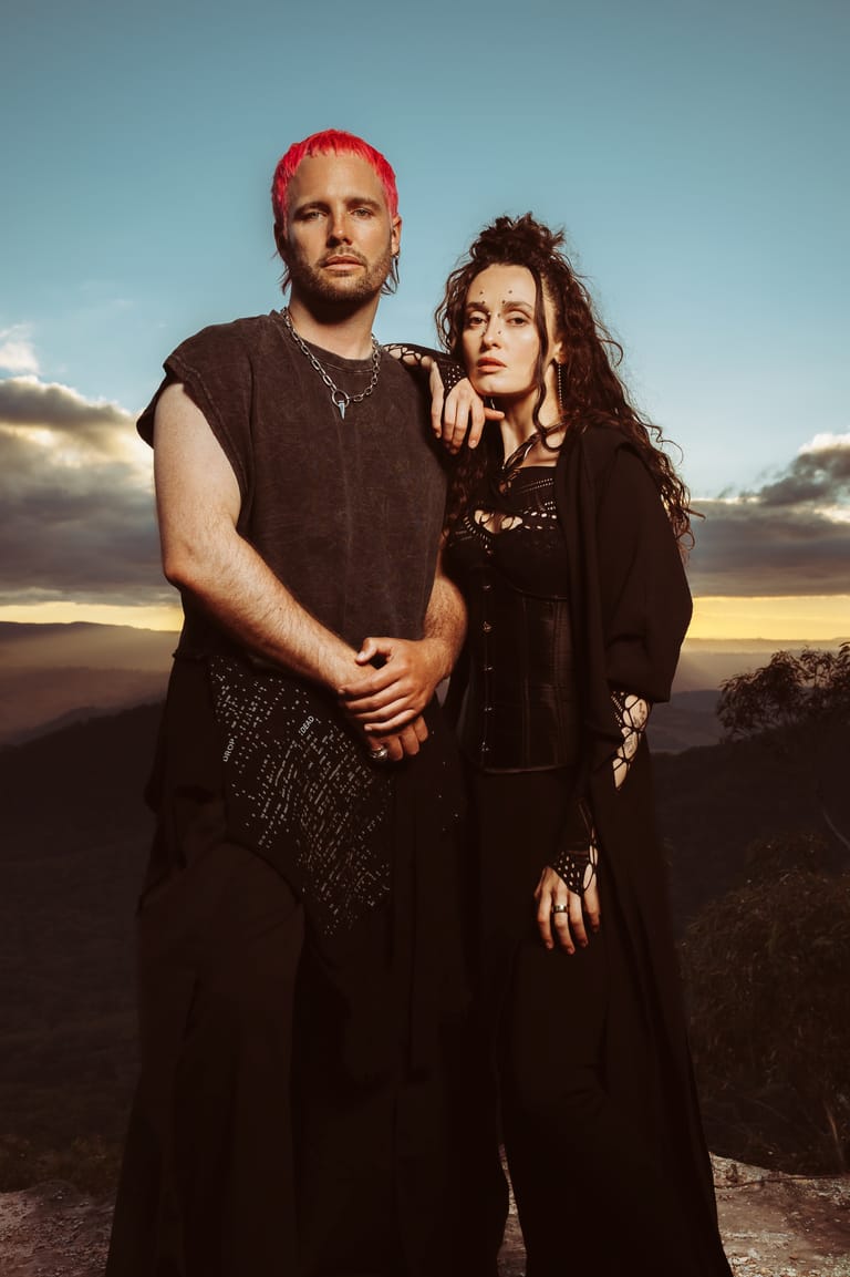Two musicians in dark clothing stand against a dramatic sunset sky, the man with bright pink hair and the woman with long dark hair.