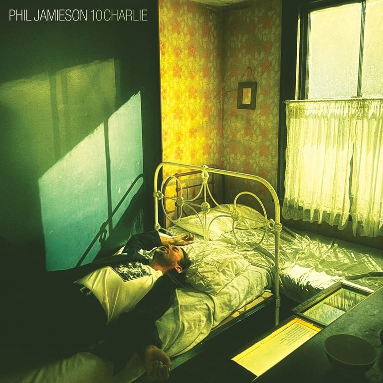 A figure lies on a metal bed in a sparse, dimly lit room with yellow-green tones and minimal furnishings.