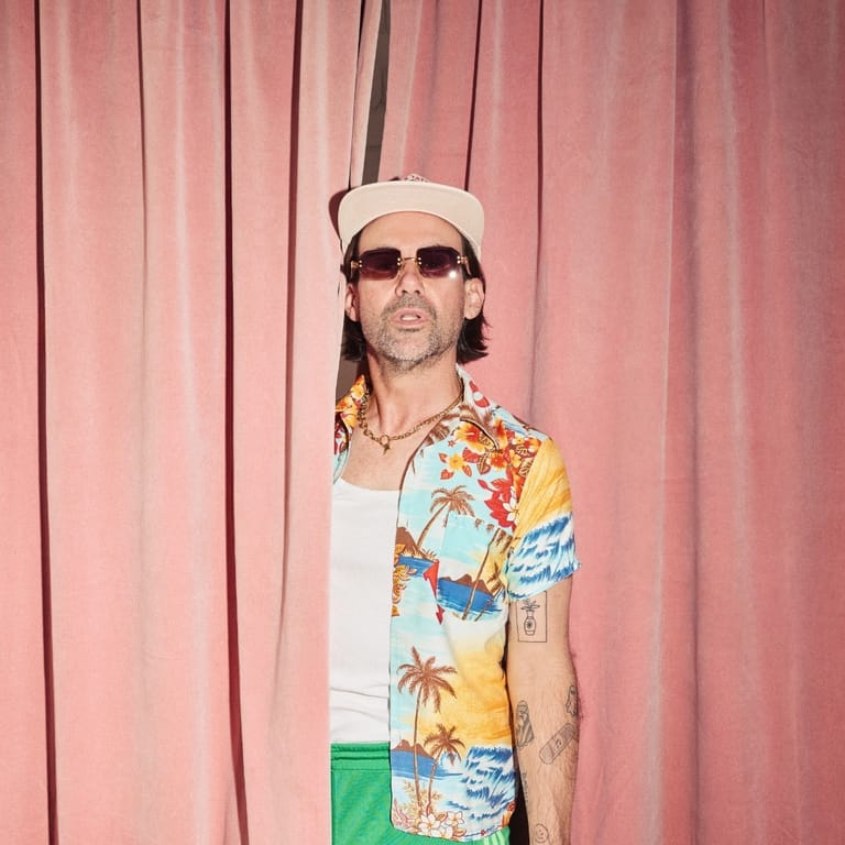 Musician in sunglasses and white cap wearing a colourful tropical-print shirt against a pink curtained backdrop.