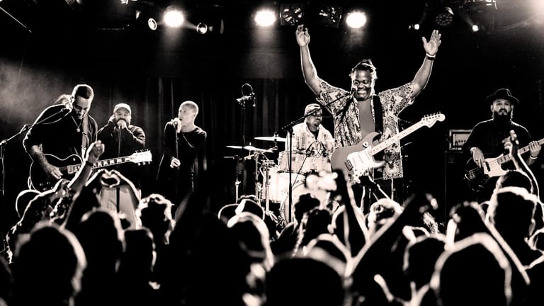 Rock band performing on stage with raised arms amid enthusiastic crowd and dramatic stage lighting.