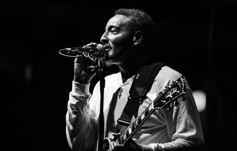 Musician singing into microphone whilst playing guitar on stage, lit by dramatic black and white lighting.