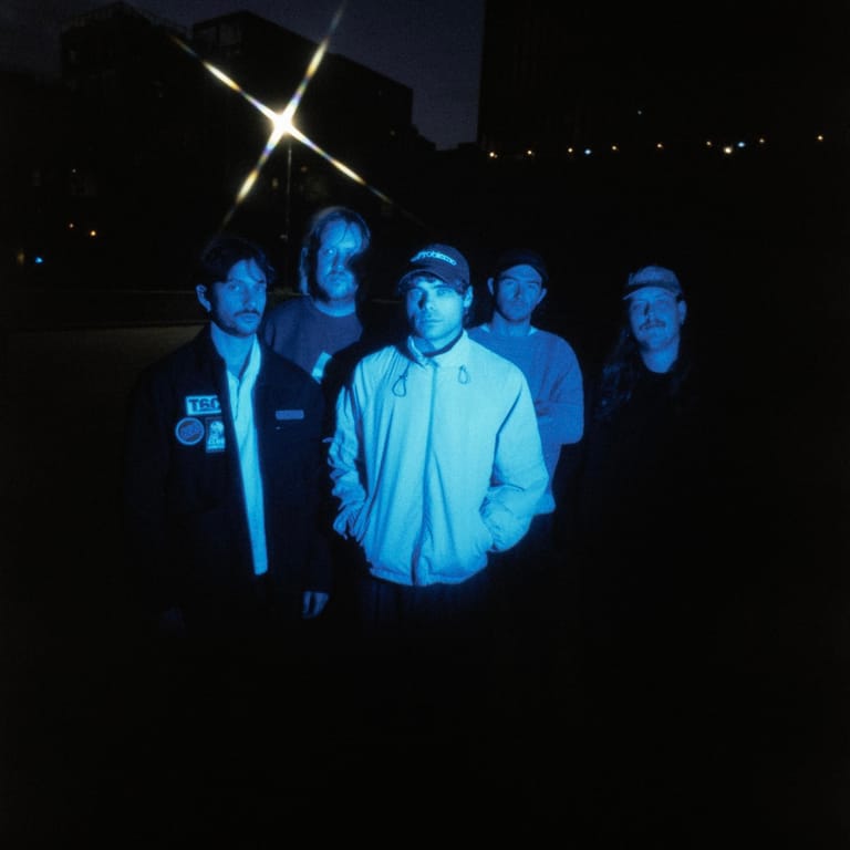 Five musicians stand in blue stage lighting against a dark night sky, with a glowing cross visible above.