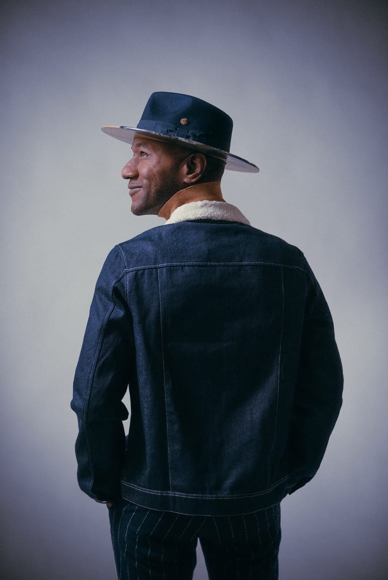 Man in navy denim jacket and wide-brimmed hat gazes over shoulder against grey background.