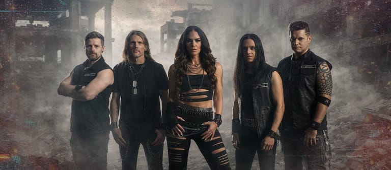 Five band members in dark clothing stand against a smoky, industrial backdrop.