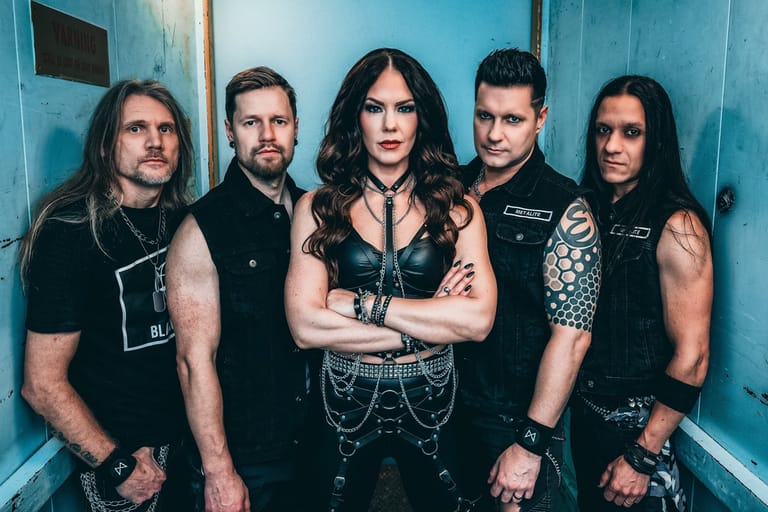 Five band members in black clothing stand against a blue wall in a studio setting.