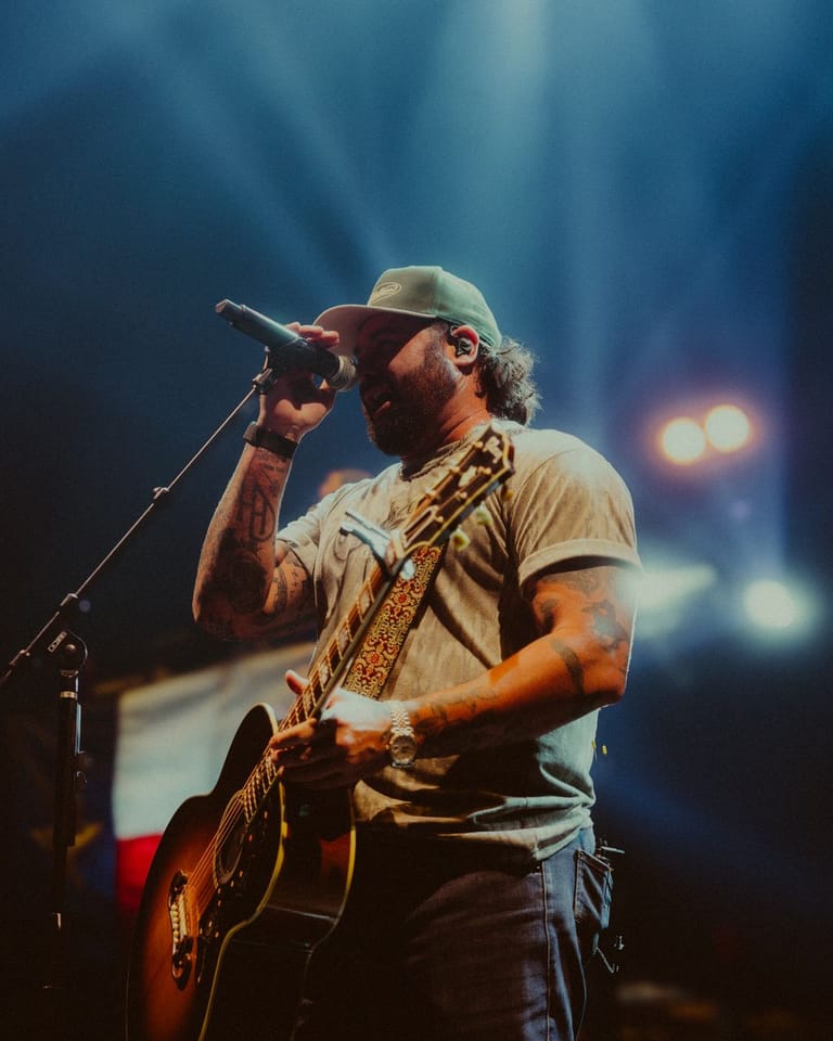 Musician in cap and tan shirt singing at microphone while playing acoustic guitar on stage.