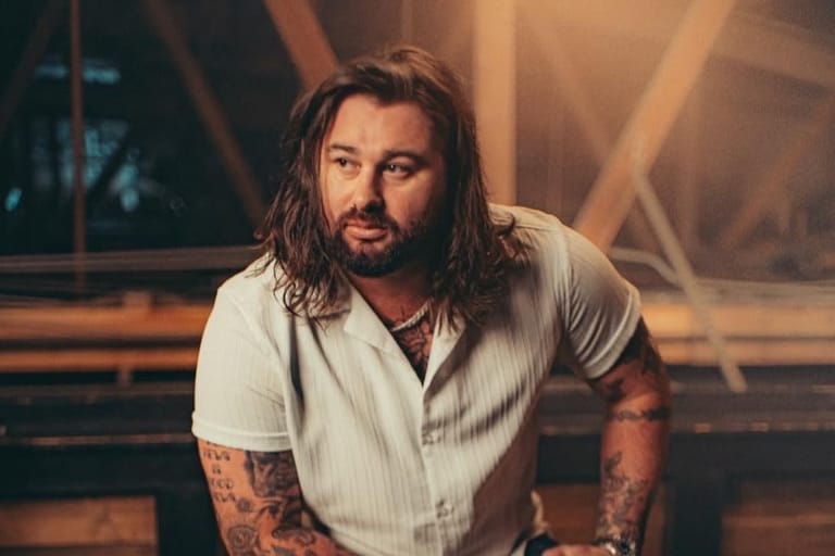 Musician with long dark hair and tattoos in white shirt against warm-lit stage backdrop.