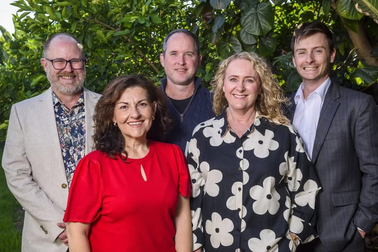 Five people pose together against a green leafy backdrop, dressed in smart casual attire.