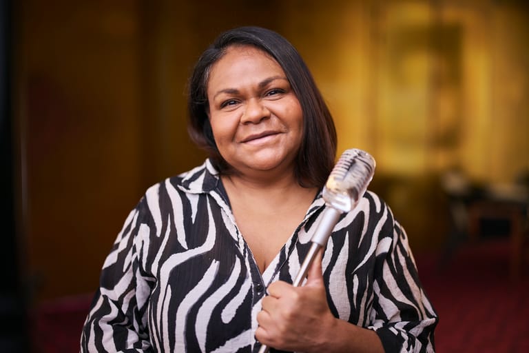 Woman in a zebra-striped shirt holding a microphone against a warm, blurred interior background.