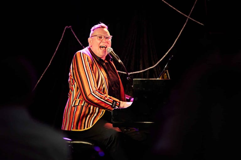 Musician in a gold-striped jacket performing at a piano under stage spotlights.