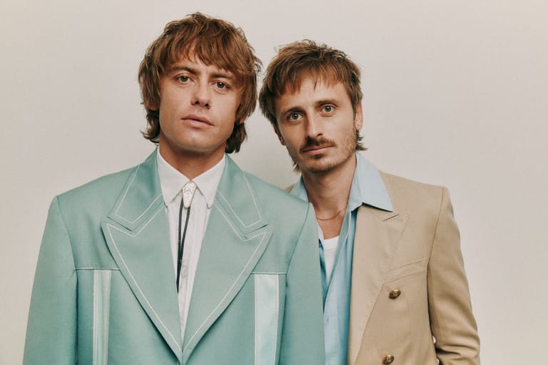 Two men in vintage-style blazers, one mint green and one beige, stand against a neutral backdrop.