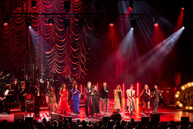 Ensemble of performers in formal attire on stage beneath red velvet curtains, bathed in dramatic red and white spotlight lighting.