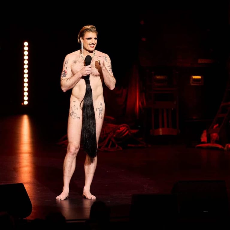 Performer in a pale patterned suit holding a microphone on a darkened stage lit by warm amber spotlights.