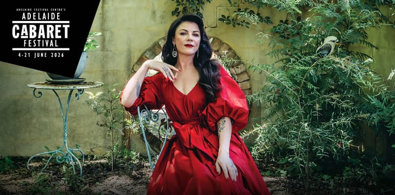 Performer in a red dress posed against a weathered green wall with foliage.