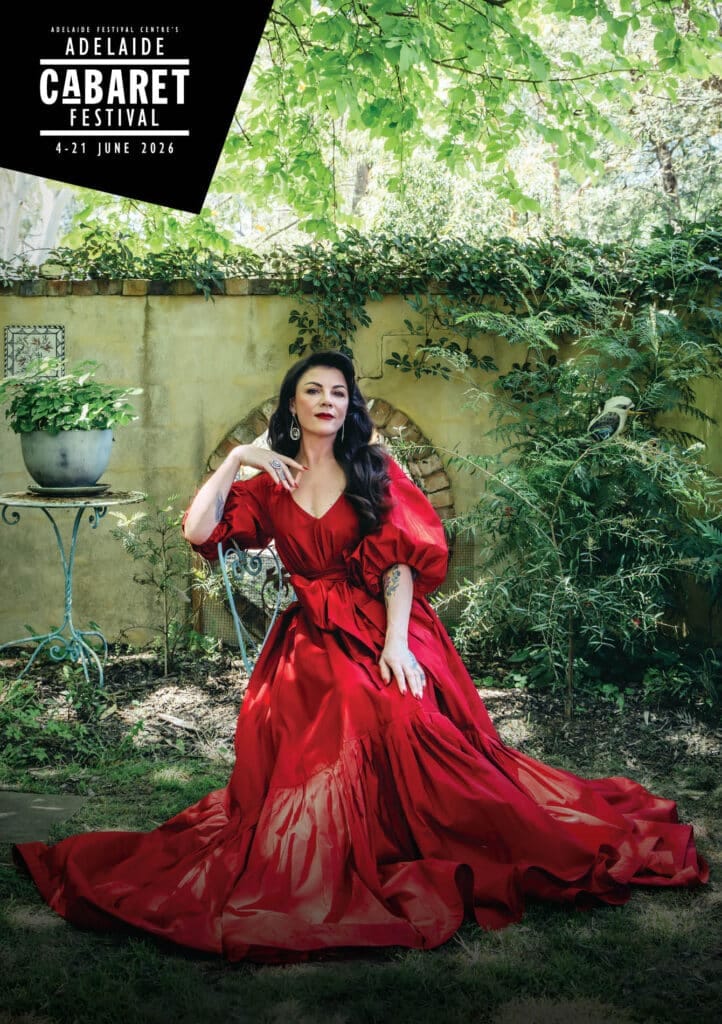 Performer in a flowing red gown poses against a weathered wall draped with climbing green ivy.