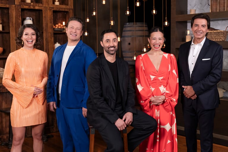 Five elegantly dressed people stand together in a warmly lit interior space with hanging pendant lights and wooden panelling.