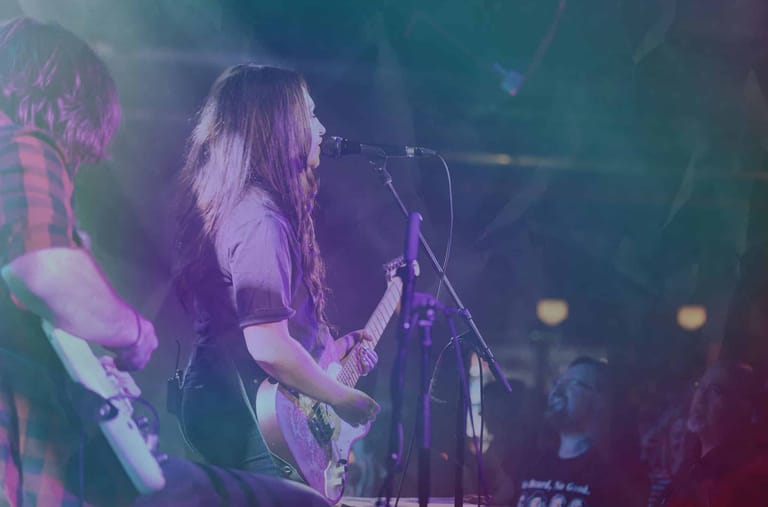 Musician with long hair performing at a microphone under purple and green stage lighting.