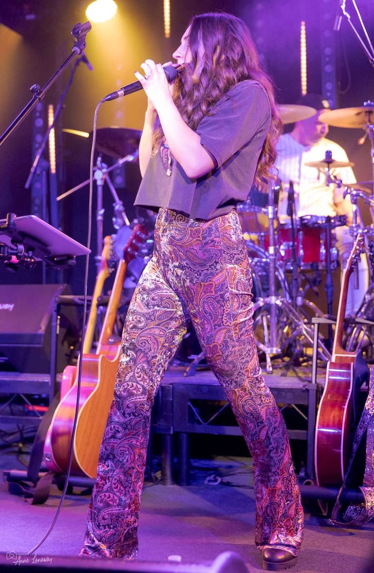 Singer performing at a microphone in patterned trousers, illuminated by purple and yellow stage lighting with band members visible behind.