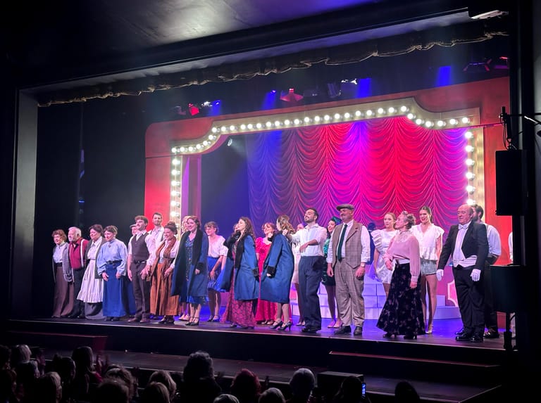 Theatre cast takes a bow on stage under vibrant pink and blue lighting during Funny Girl production.