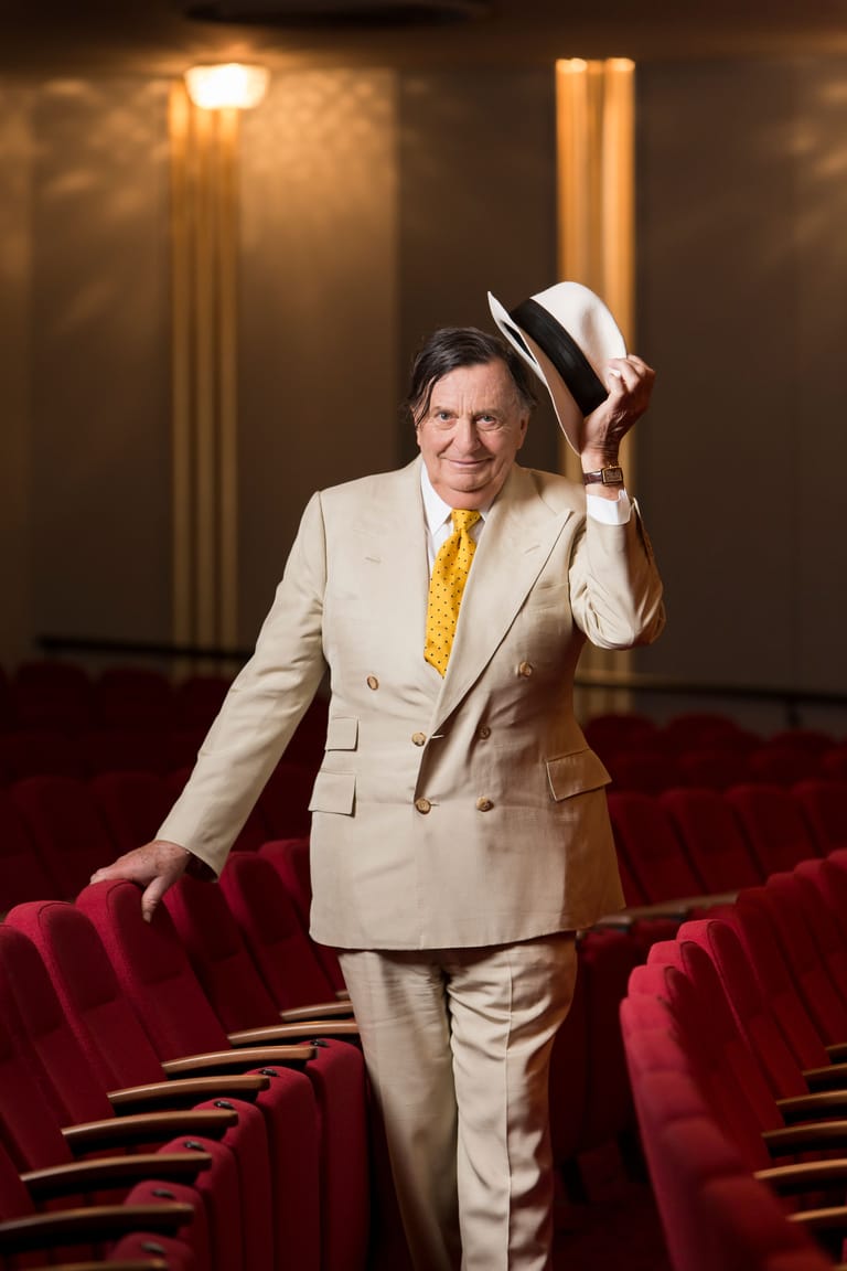 Man in cream double-breasted suit and panama hat stands between red theatre seats, tipping his hat.