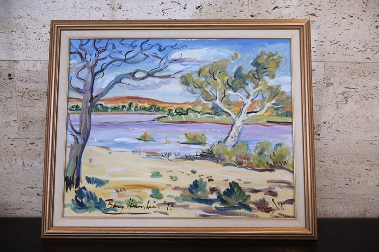 Framed landscape painting depicting a river scene with bare and leafy trees, sandy banks, and distant hills under a blue sky.