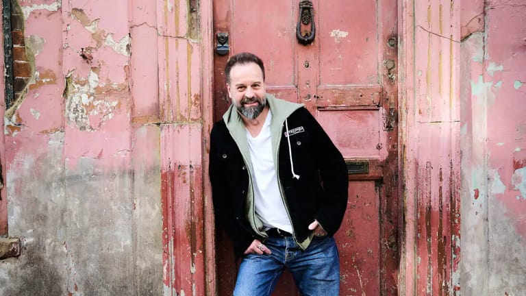 Man in black jacket and jeans leaning against weathered pink and red doors.