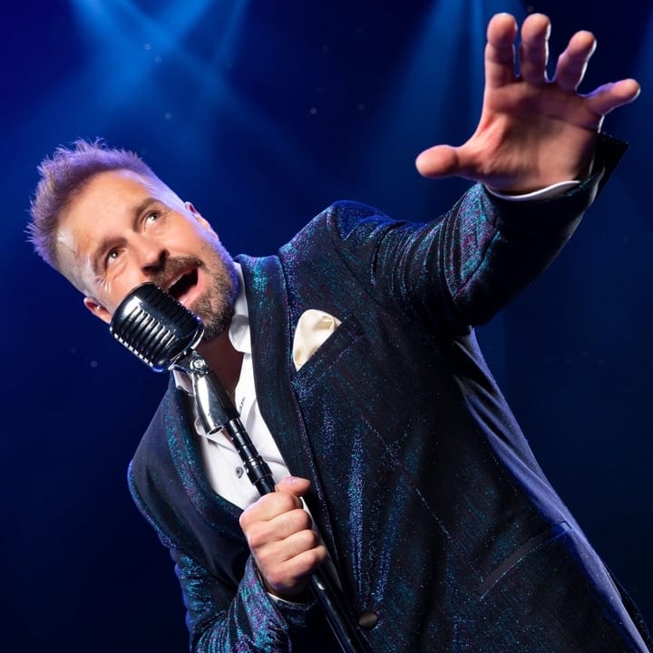 Male performer in a dark blue jacket singing into a vintage microphone with arm extended against blue stage lighting.
