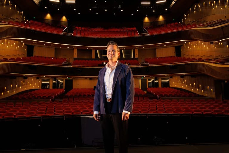 Person standing centre stage in a theatre with red seating and warm overhead lighting.