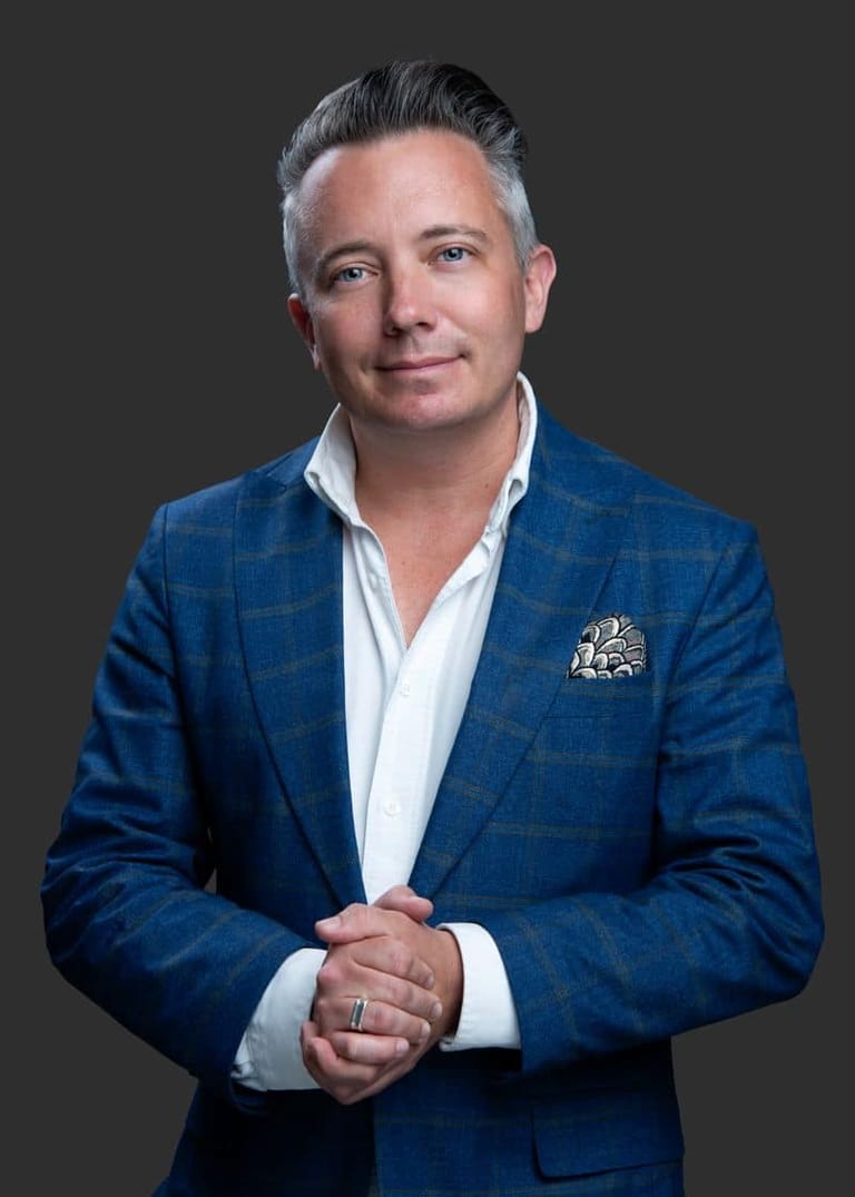 Man in blue checked blazer and white shirt against neutral grey background, hands clasped at chest.