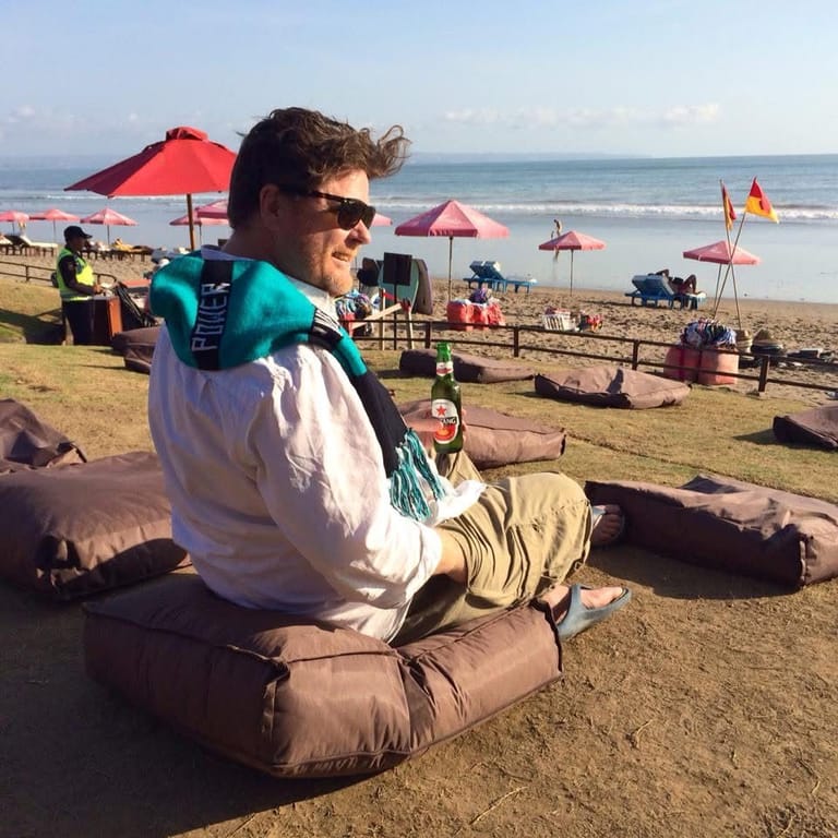Man in sunglasses and casual beachwear relaxing on sand with red umbrellas and beachgoers visible behind.
