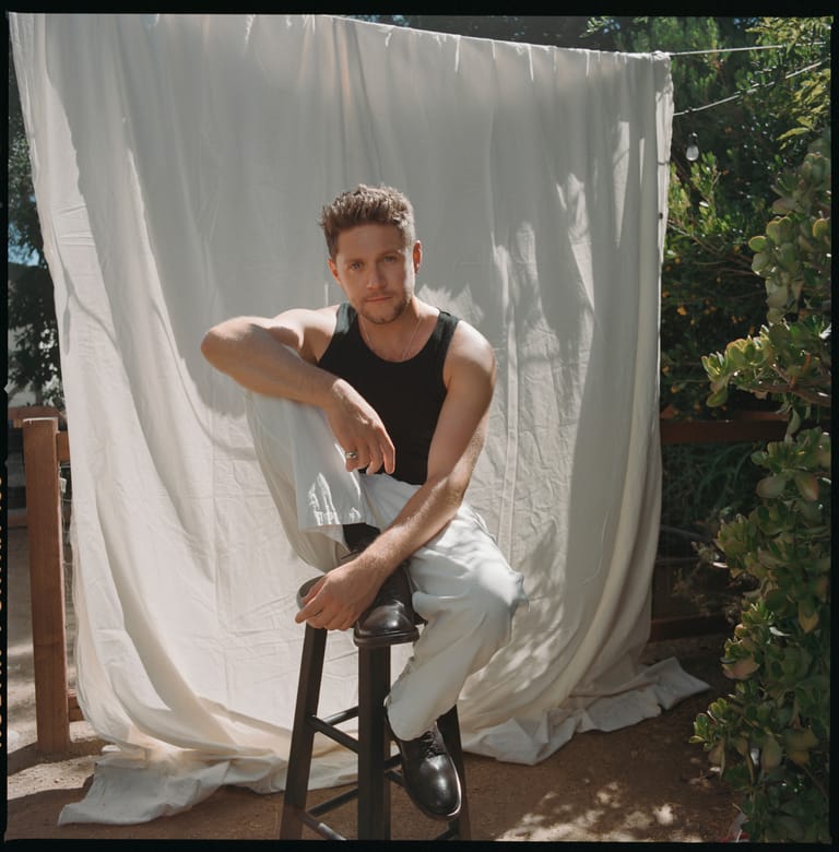 Person in black sleeveless shirt seated on a stool against a white fabric backdrop outdoors.