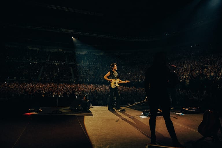 Musician in yellow shirt playing guitar on stage before a large crowd in a darkened venue.