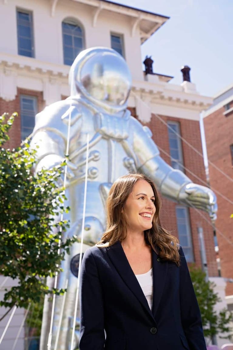 Woman in dark blazer smiles beside large illuminated astronaut sculpture outside historic brick building.
