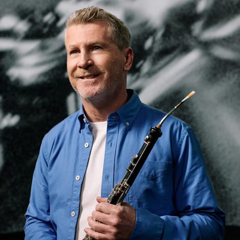 Man in blue shirt holding an oboe against a blurred grey background.