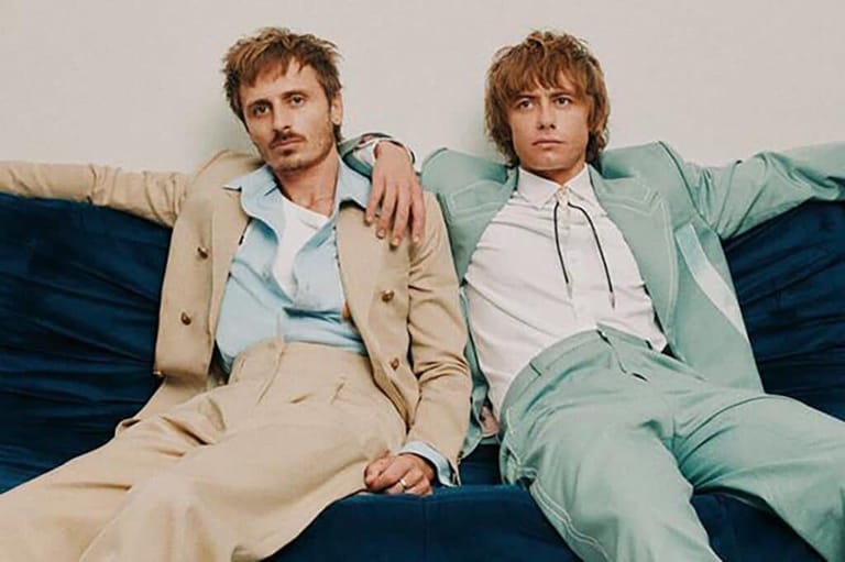 Two men in coordinating pastel suits recline on a blue sofa against a neutral background.